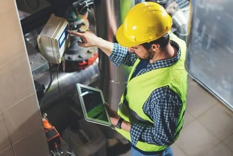 A man wearing a hard hat and safety vest is focused on using a tablet in a construction environment.
