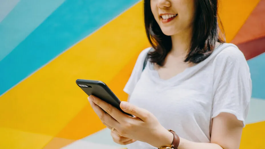 A woman smiles brightly while holding a cell phone in her hand, exuding joy and positivity.