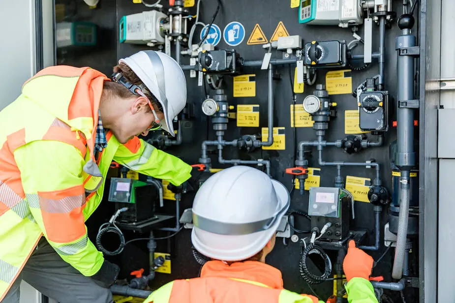 Two men in safety vests are collaborating on a panel, focused on their task in a work environment.