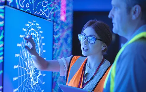 A man and woman in safety vests observe a large screen, engaged in a discussion or analysis of displayed technical information.